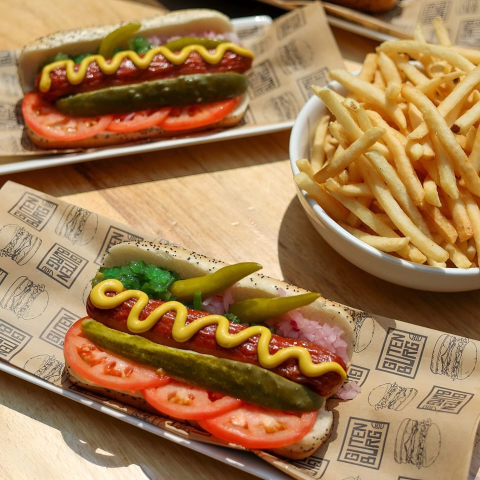 Two Gutenburg hot dogs with mustard, relish, pickles, and tomatoes, served with a side of fries.