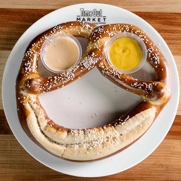 A large, salted pretzel with two dipping sauces, one yellow mustard and one light brown, on a white plate.
