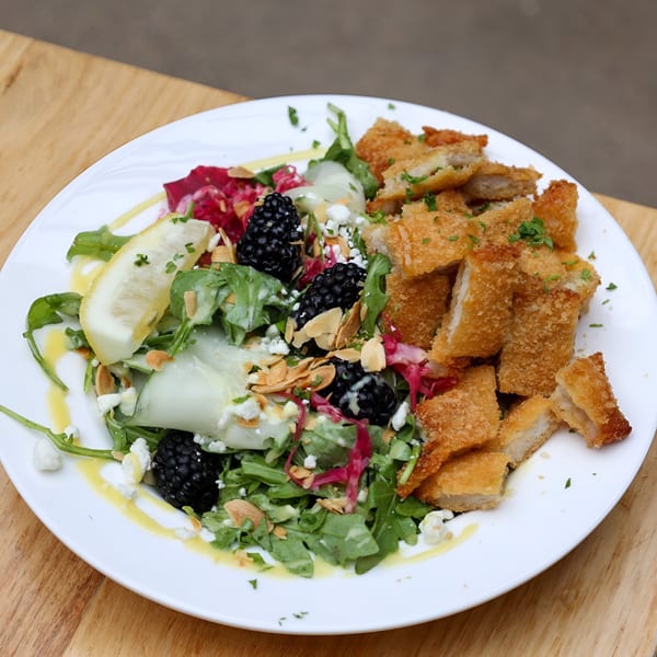Crispy fried chicken pieces with a fresh salad, blackberries, feta, and lemon on a white plate.